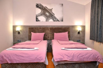 Хотелска стая с две единични легла, розово спално бельо и модерно осветление.